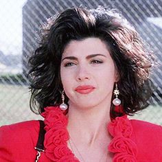 a woman in a red shirt and earrings standing next to a chain link fence with her hands on her hips