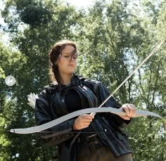 a woman in black jacket holding a bow and arrow with trees in the back ground