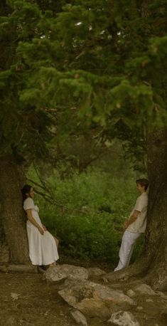 two people in white dress standing next to each other near trees and rocks on the ground