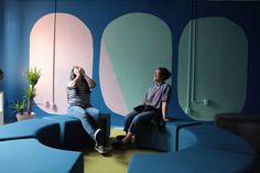 two women sitting on blue couches in front of a colorful wall