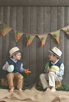 two young boys are sitting on hay with carrots and vegetables in front of them