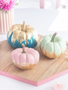 three painted pumpkins sitting on top of a wooden tray next to a pink vase