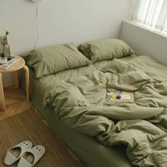 an unmade bed with green sheets and slippers on the floor next to it