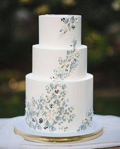 a three tiered white wedding cake with blue flowers on the side and gold trim