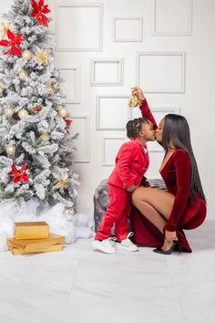 a woman kneeling down next to a small child in front of a christmas tree with presents