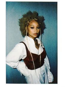 a woman with curly hair wearing a white shirt and brown suspenders, standing in front of a blue wall