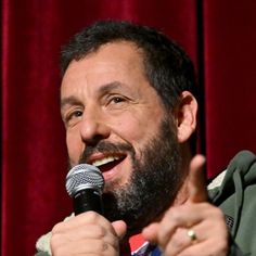 a man with a beard holding a microphone in front of a red curtain and pointing to the side