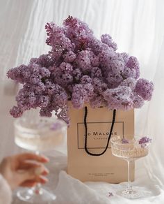 purple flowers are in a paper bag and someone is holding a wine glass next to it