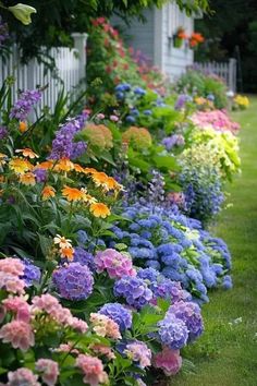 many different colored flowers line the side of a house with white picket fence in the background