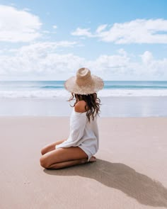 a woman sitting on the beach wearing a hat