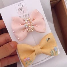 two small bows with pearls on them are in a box, one is pink and the other is yellow