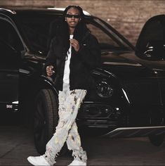 a man standing in front of a black car wearing white sneakers and an oversize jacket