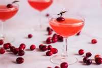 cranberry martinis in coupe glasses with cherries scattered around them on a white surface