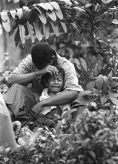 two people sitting on the ground in front of some plants and trees, one is hugging the other's head