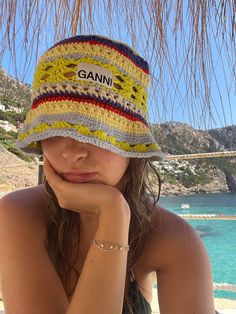 a woman wearing a hat sitting on top of a beach next to the ocean with her hand under her chin