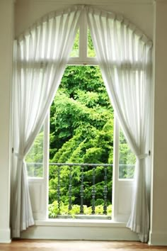 an open window with white curtains and green trees in the background, looking out onto a wooden floor