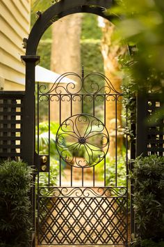 an iron gate in front of a house with trees and bushes around it, leading into the yard