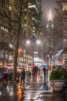 people walking down the street in the rain at night with umbrellas and buildings behind them