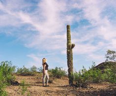 a woman standing next to a tall saguado cactus on top of a dirt field