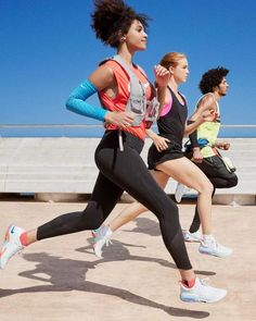 three women are running on the beach in their sports clothes and shoes, one is wearing an orange tank top