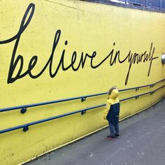 a little boy standing in front of a yellow wall with writing on it that says believe anyway