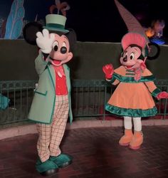 mickey and minnie mouse in costumes standing on a brick walkway at night with lights behind them