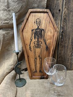 a wooden box with a skeleton on it next to two wine glasses and a candle