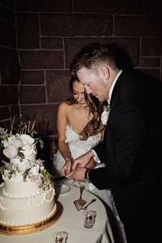 a bride and groom cutting their wedding cake at the reception table in front of brick wall