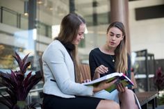 two women sitting on a bench looking at a book while one woman is holding a folder