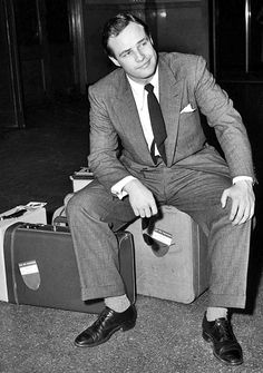 a man in a suit and tie sitting on a piece of luggage with his legs crossed