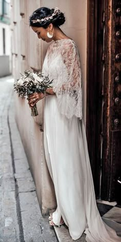 a woman standing next to a building holding a bouquet of flowers and wearing a white dress