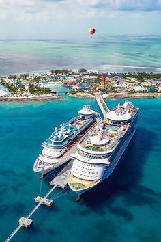 two cruise ships docked in the water near an island and some buildings with hot air balloons flying over them