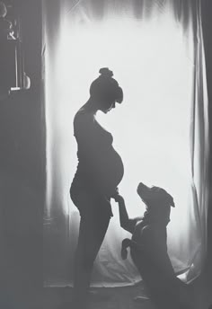 a pregnant woman holding her dog's hand while standing in front of a window