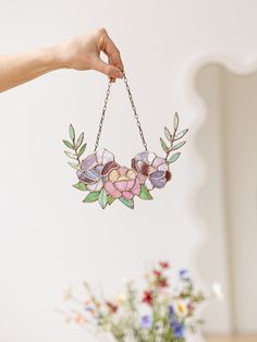 a hand holding a stained glass hanging decoration with flowers and leaves on the chain, in front of a vase filled with wildflowers