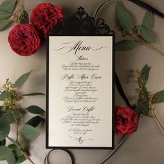 a wedding menu with red roses and greenery on the table next to it,