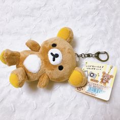 a brown teddy bear keychain laying on top of a white bed next to a tag