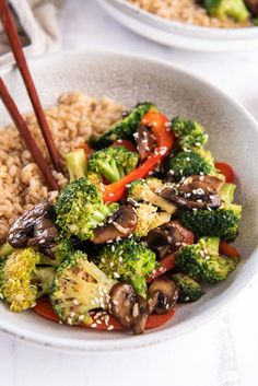 a white bowl filled with broccoli, mushrooms and other veggies next to brown rice