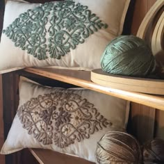 three decorative pillows and two balls of yarn on a wooden shelf next to a ball of yarn