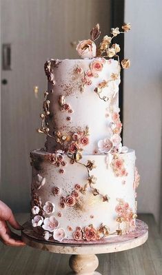 a three tiered wedding cake with flowers on the top is being held by a person