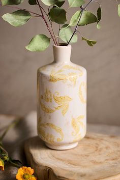 a white vase with yellow flowers in it on a wooden table next to some green leaves