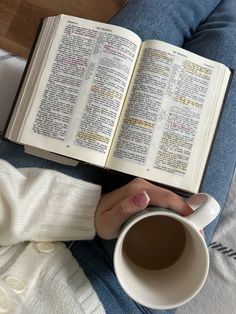 a person sitting on a bed holding an open book and cup of coffee in their hands