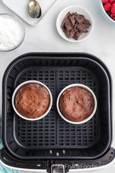 two chocolate muffins sitting on top of an air fryer next to bowls of raspberries