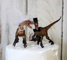 two toy dinosaurs on top of a white cake with a bride's dress and groom's hat