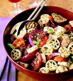 a bowl filled with pasta and vegetables on top of a wooden table next to a glass of wine