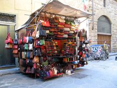 an outdoor market selling handbags and purses on the side of a street in front of a brick building