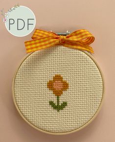 a cross stitch flower with a yellow bow on it's head is displayed in front of a pink background