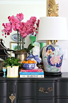 a black dresser topped with lots of books and vases filled with pink flowers next to a lamp