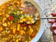 a person holding a spoon over a bowl of soup with vegetables and herbs in it