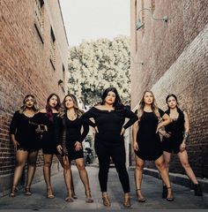 a group of women standing next to each other in front of a brick wall on a street