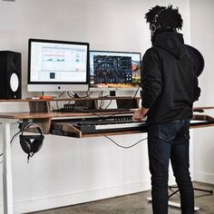 a person in a black hoodie standing at a desk with two computer monitors and keyboards